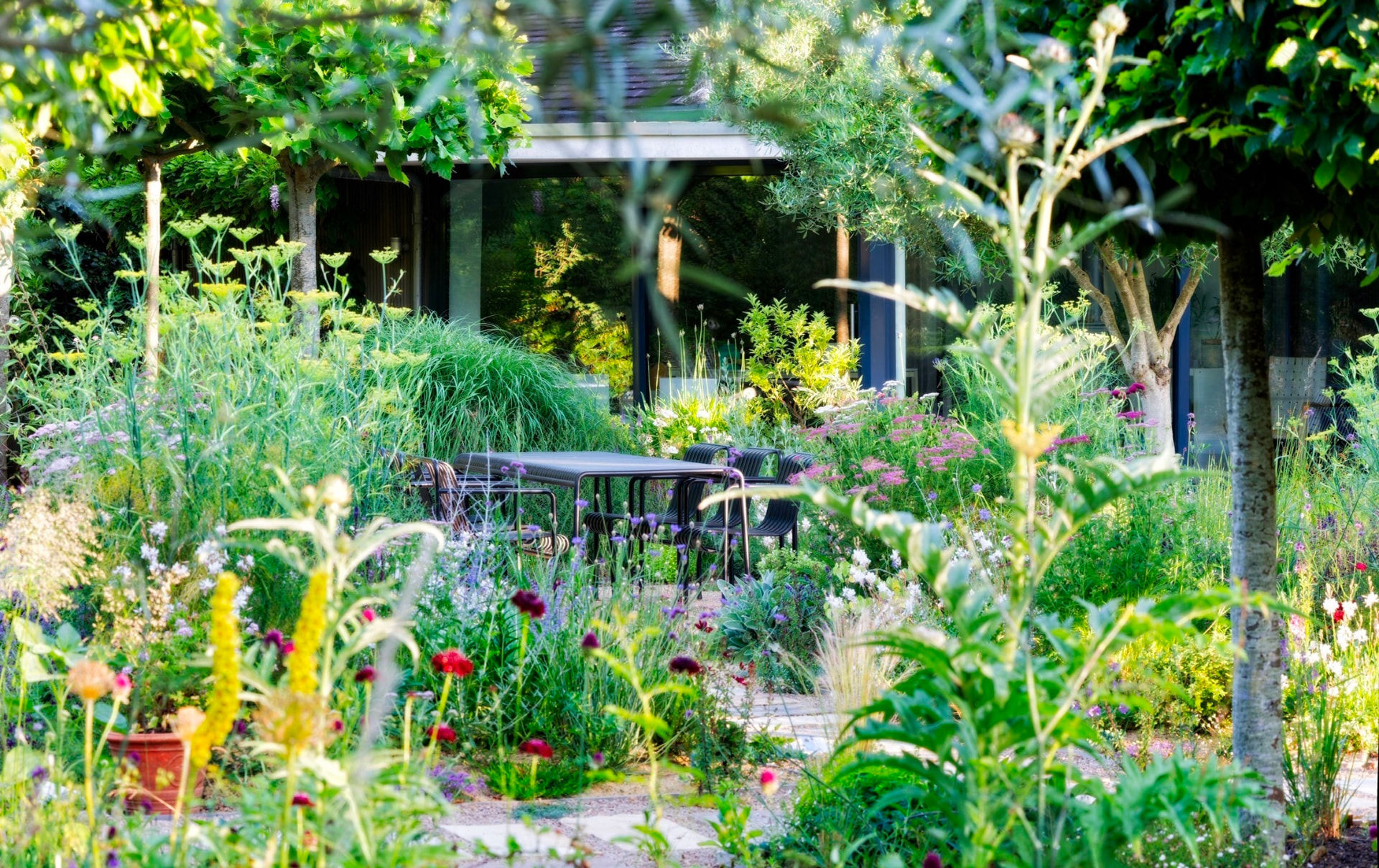 A lush Mediterranean gravel garden with a black dining table, tall grasses, and purple verbena